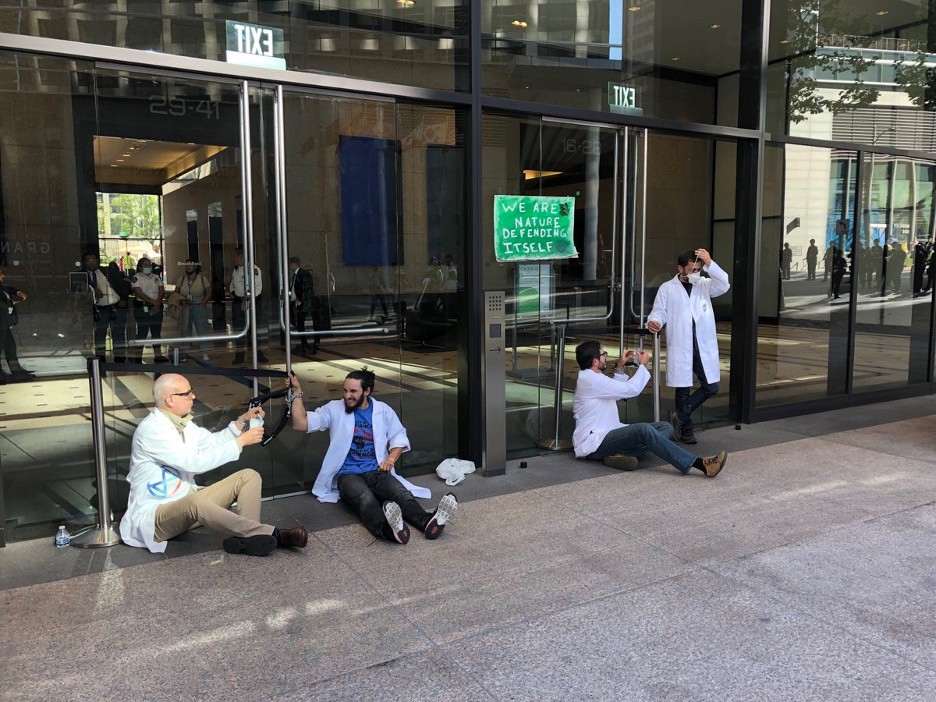 Scientist Rebellion action at Chase Bank Image: Four scientists dressed in white lab coats sit on the ground, with their hands chained to large glass doors outside Chase bank